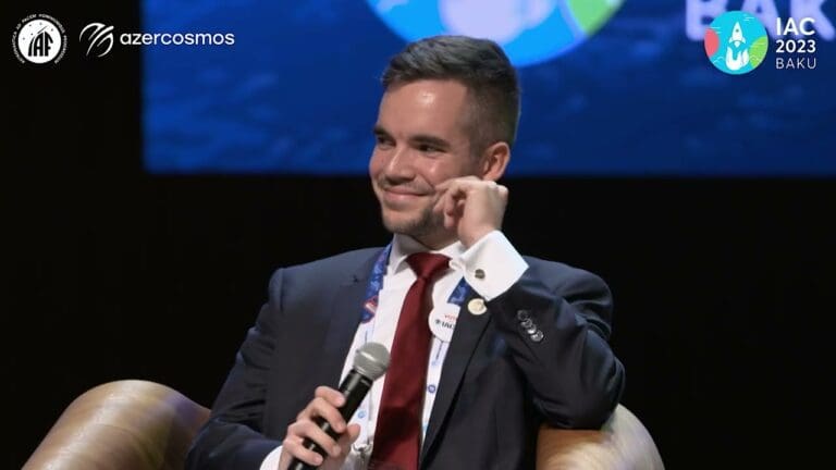 A smiling man in a suit sits on stage holding a microphone at a panel discussion on Space Exploration at IAC 2023 Baku, with Azercosmos and IAF logos in the background